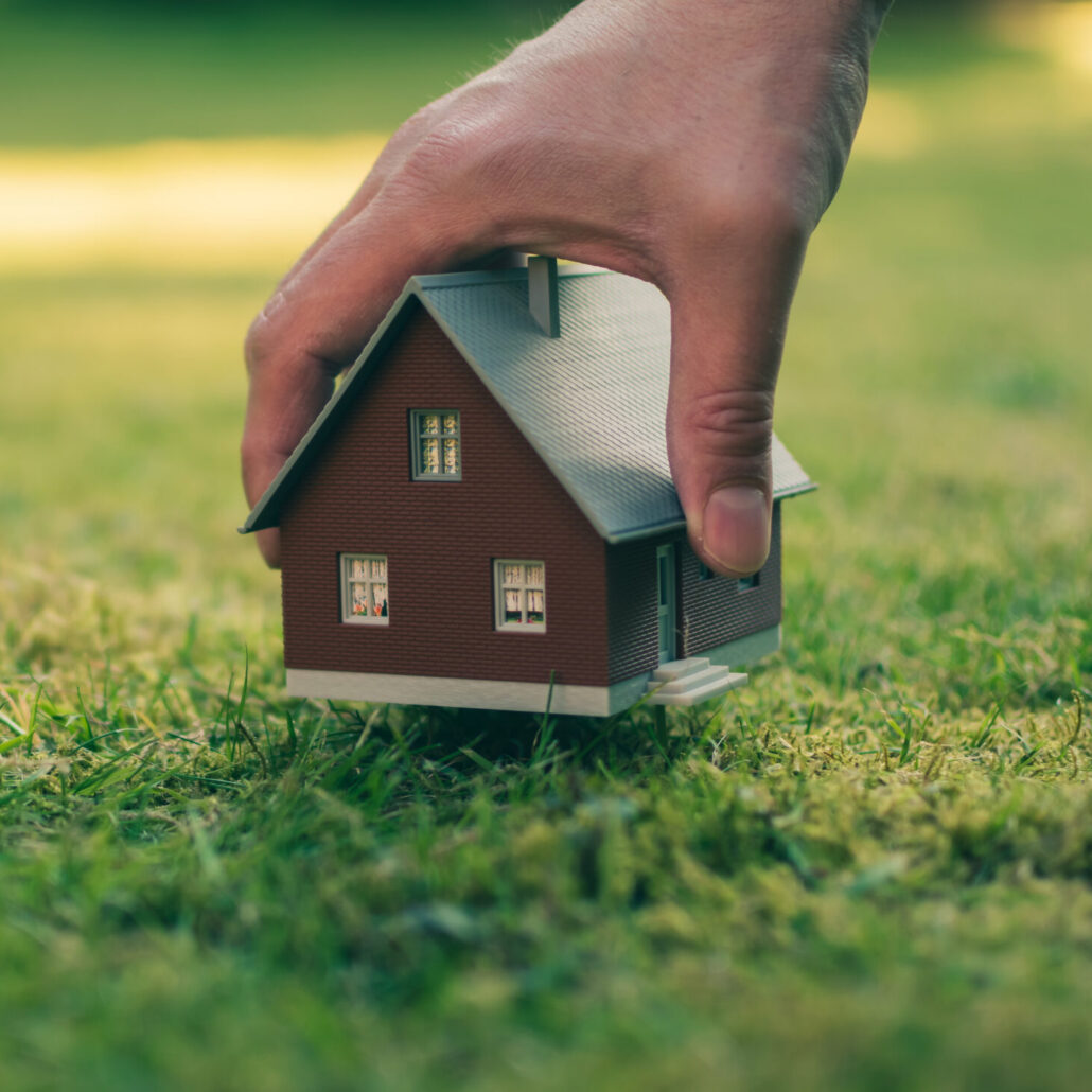 concept of selling a house. a hand is holding a model house above green meadow.